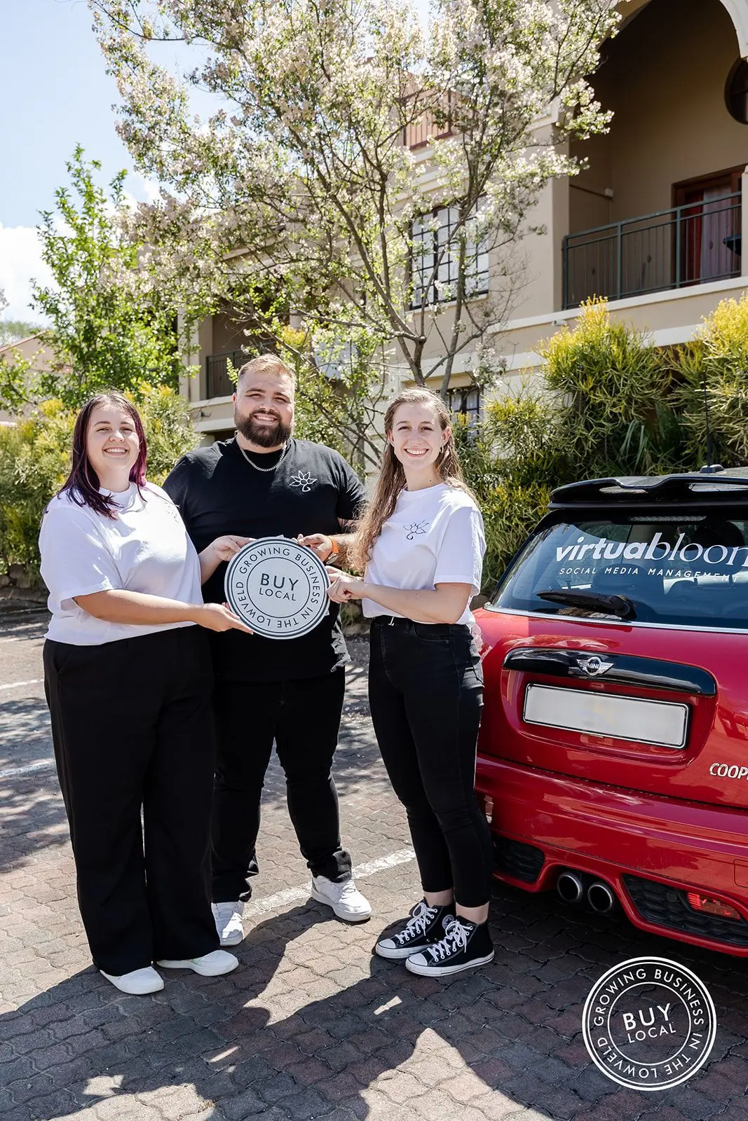 Tammy, Shandon, and Melissa standing next to a red car holding the Buy Local Lowveld logo